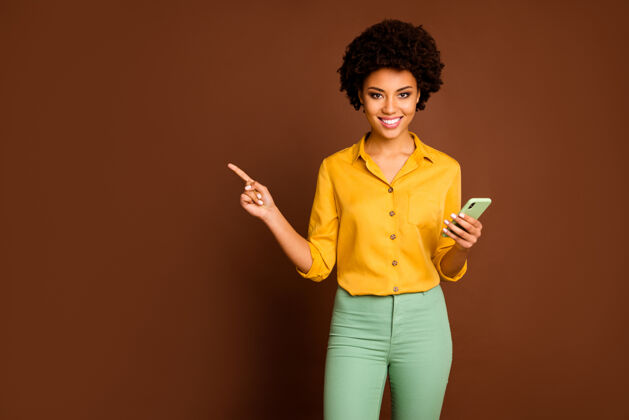 衬衫美丽的深色皮肤波浪女士手持电话的照片影响者表示手指空位显示销售应用程序价格穿黄色衬衫绿色裤子孤立棕色信息指向混合空间