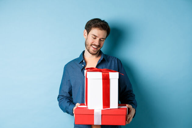 丈夫浪漫的年轻人 留着胡子 看着情人节的礼盒 给爱人送礼物 站在蓝色的背景上微笑男人温柔