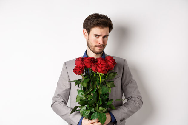 日子犹豫不决的留着胡子的男人穿着西装 拿着鲜花和玫瑰 看着相机不确定 去浪漫的约会 站在白色的背景上惊喜同性恋情人节