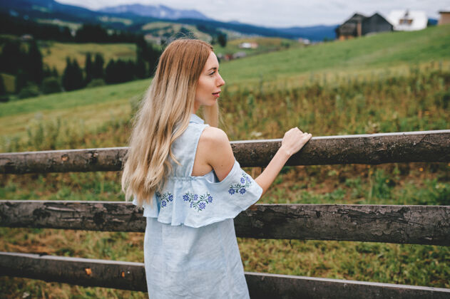 木头年轻漂亮的金发碧眼的女孩穿着蓝色的裙子 在乡村的篱笆旁摆姿势乡村如画女孩