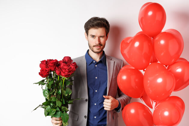 情人自信的男朋友穿着西装去浪漫的约会 对着镜头眨眼 捧着一束红玫瑰 站在心气球旁边眨眼 站在白色背景上西装情人节花