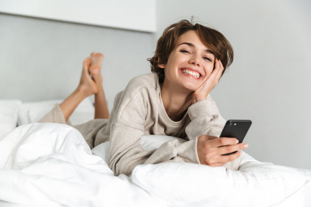 女人微笑的年轻女孩 早上在床上放松 用手机年轻连接手机