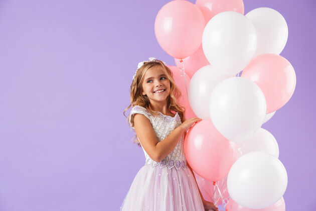 肖像可爱漂亮的小女孩穿着公主裙 隔着紫罗兰色的墙 手里拿着一堆气球束童年爱