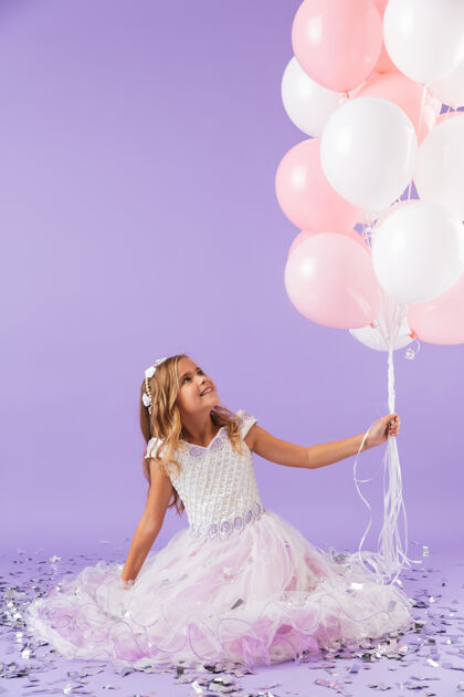 小穿着公主裙的漂亮小女孩孤立地坐在紫罗兰色的墙上 手里拿着一堆气球空气五颜六色年轻