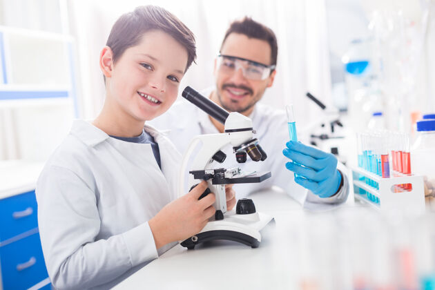 实验室最好的作品热情快乐的男孩一边看着相机一边穿着实验服调显微镜年轻分析技能