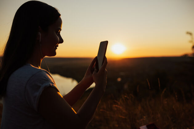 年轻漂亮女孩在看电话日落结束-在日落的背景下 一个女孩拿着智能手机美丽风景天空