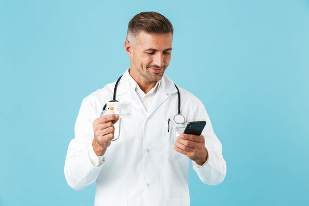 电话中年医生身穿白大褂 手持听诊器手持智能手机 孤零零地站在蓝色墙壁上的照片男人医生治疗