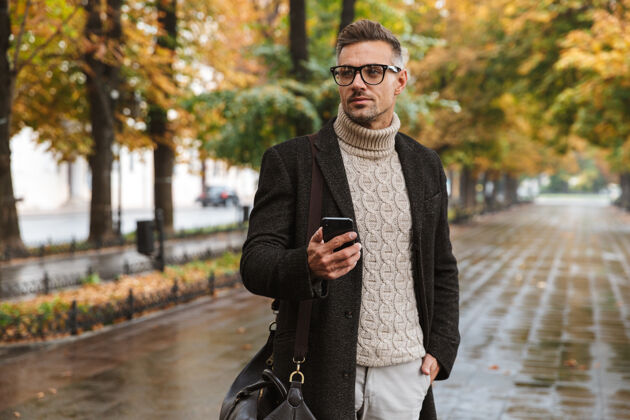 帅气一张30多岁的留胡子男人穿着暖和的衣服在户外穿过秋天公园 并使用智能手机的照片公园吸引力空旷