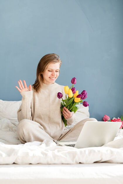 快乐快乐微笑的女人穿着睡衣坐在床上 愉快地赏花 工作在笔记本电脑上日子卧室房子
