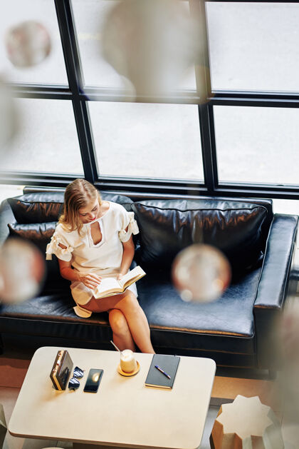 卡布奇诺美丽的金发女郎坐在皮沙发上读着有趣的书 从上面看女性休闲活动坐着