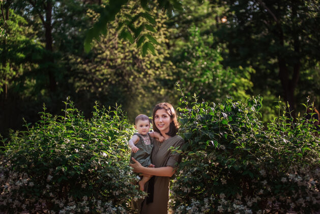 享受一个年轻的女人怀里抱着一个小婴儿一位美丽的母亲和她的女儿在茉莉花丛旁的绿色公园里散步可爱公园自然