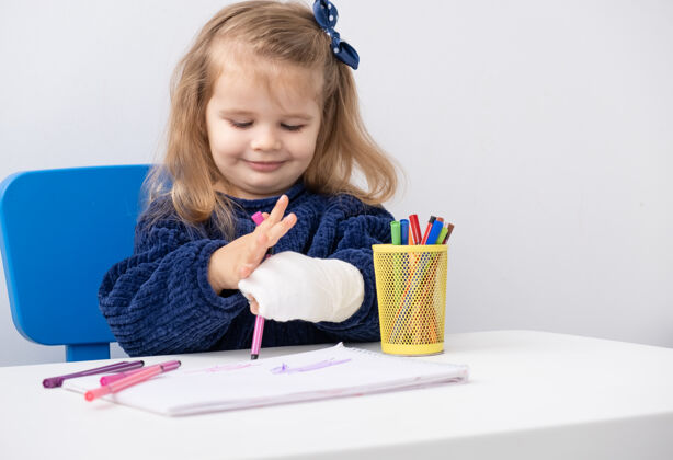 矫形坐在桌边用马克笔试着画画的小女孩手拿石膏手事故医学
