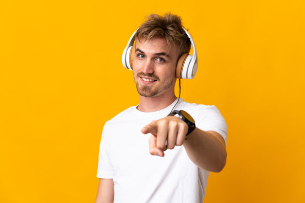 科技年轻英俊的金发男人孤立在黄色背景上听音乐听力男人电话
