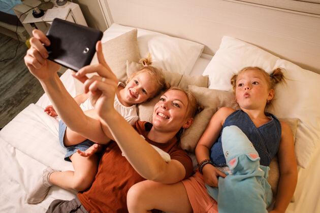 自拍妈妈和她的两个女儿在床上休息的时候用智能手机自拍放松兴奋缩放