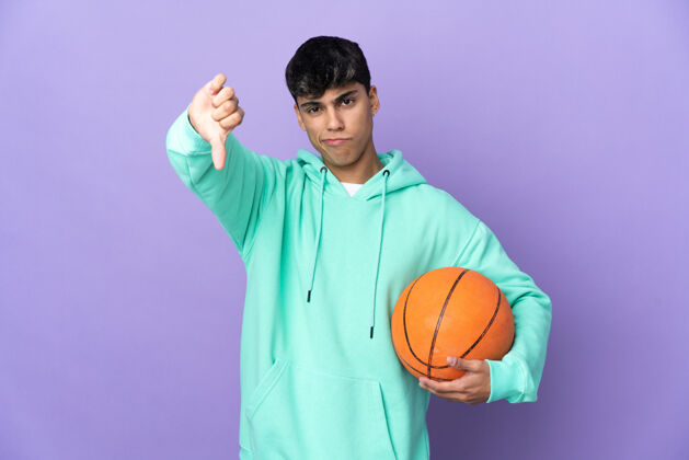 不喜欢在孤立的紫色背景上打篮球的年轻人用否定的表情展示拇指朝下运动装制服减肥