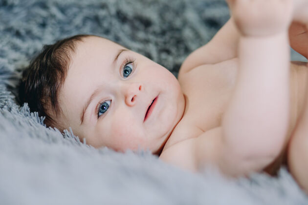 护理六个月大的裸体婴儿躺在柔软的灰色床单上 抱着她的脚趾欢呼小房间