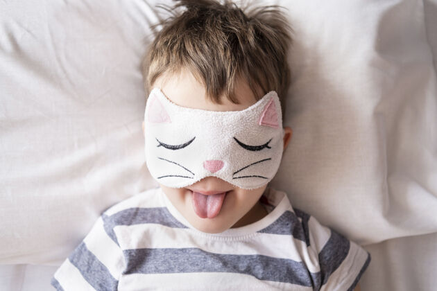 醒来白人有趣的学龄前男孩在小猫眼罩条纹睡衣躺在白色的床上白种人幼儿园孩子