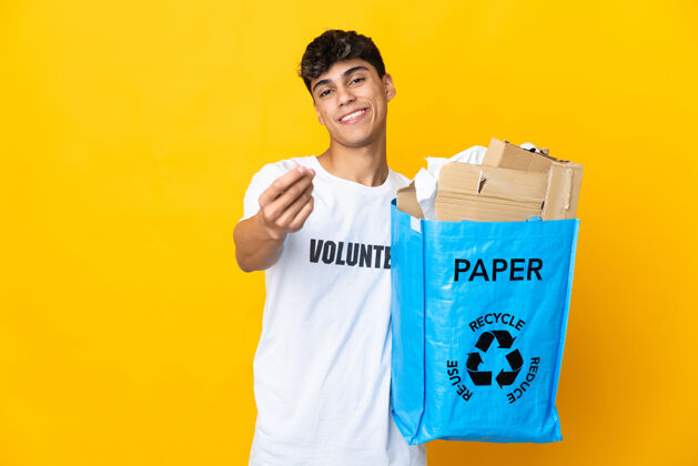 年轻男人拿着一个装满废纸的回收袋 在孤立的黄色背景下进行回收赚钱的手势男性财政环境