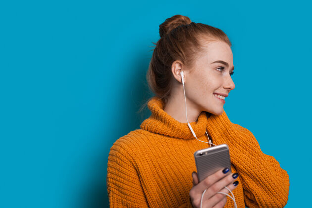手机满脸雀斑 满头红发的白种女人微笑着 一边看着某处 一边拿着带耳机的手机 站在一堵蓝色的墙壁上 有空余的空间Mp3漂亮女孩