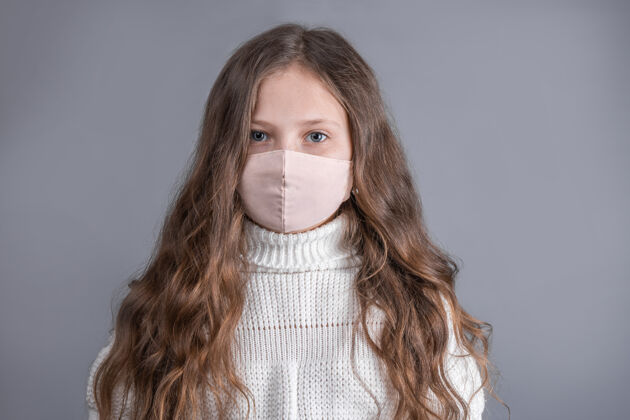 脸一个年轻漂亮的小女孩的肖像 金发飘逸 穿着白色毛衣 戴着医疗防护面具 在灰色的工作室里背景.地点为了文本.复制空间检疫疾病疾病