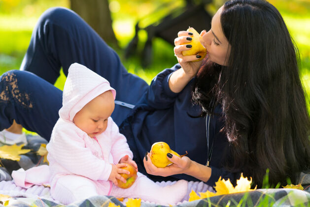 公园年轻的母亲和她的小女儿一起吃着秋天的苹果 他们一起在公园的毯子上放松爱可爱宝贝