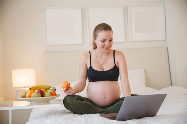 居家一个年轻的金发孕妇坐在床上 一边吃水果一边看着她笔记本电脑高高质量的照片演习水果吃