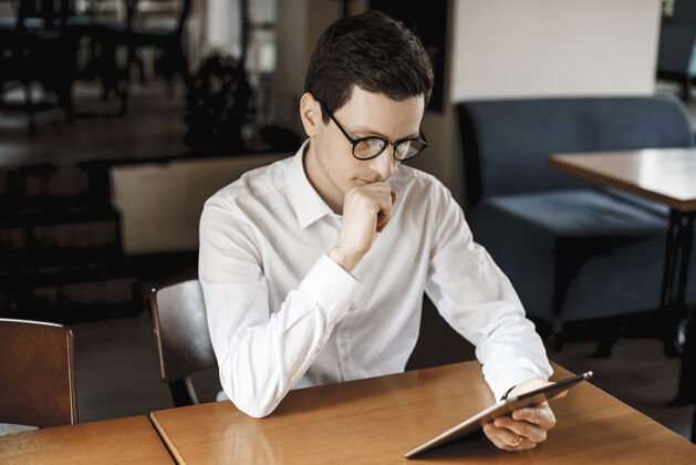 小工具穿着白衬衫 戴着眼镜 坐在书桌旁看着平板电脑的可爱年轻人休闲商务人士职业