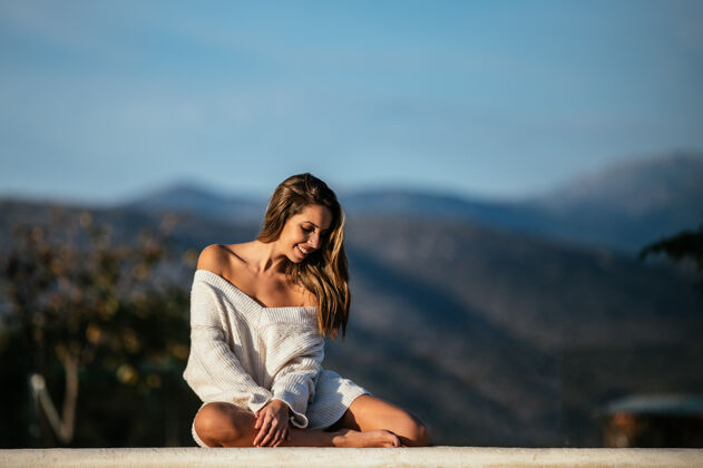 肖像穿着柔软毛衣的积极女性微笑着闭上眼睛 在模糊的山峦背景下 在院子里的边界上放松休闲毛衣微笑