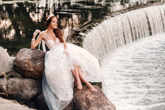 肖像一位穿着白色礼服 戴着手套 赤脚的优雅新娘正坐在公园的瀑布旁 享受着大自然美丽俄罗斯石头