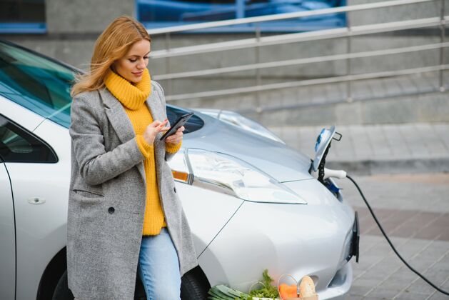 人使用智能手机等等女人在白天的电动汽车充电站轮胎尼斯汽车