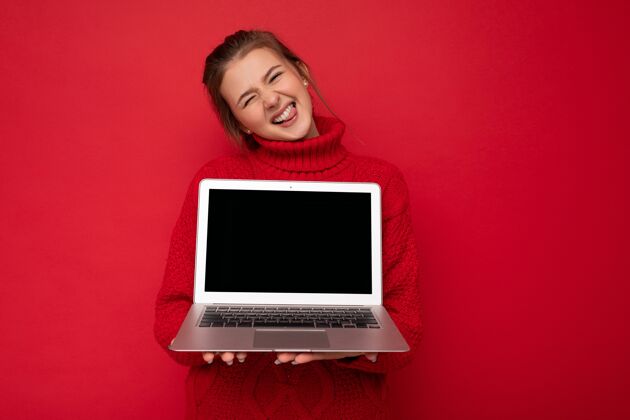 上网本美丽满意快乐的年轻女子手持电脑笔记本穿着红色毛衣展示舌头隔着红墙互联网无线电脑