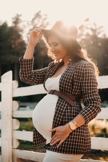 成人一个怀孕的女孩 肚子很大 戴着帽子 站在马厩附近日落时尚一个穿着棕色裙子的孕妇 骑着马帽子夜晚快乐