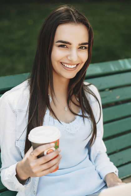 生活方式一位年轻漂亮的女性坐在长凳上 一只手拿着咖啡 看着摄像机 在城外欢笑城市大学交流