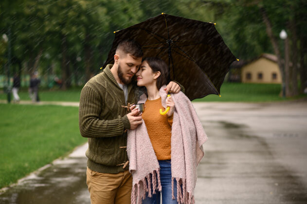 湿情侣在公园喝热咖啡 夏天下雨戴曼雨中的女人站在雨伞下 漫步小径上的浪漫约会 小巷里的潮湿天气户外女朋友快乐