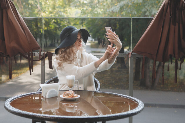 金发一位金发女郎的肖像 戴着黑色帽子 坐在咖啡馆的桌子旁 手里拿着智能手机 长发 米色外套.取下自拍成人女士杯子