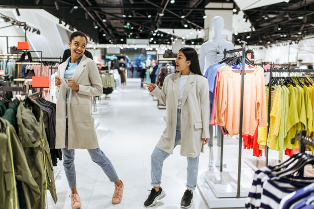 顾客两个女孩试着穿外套女店员在时装精品店购物 购物狂 在衣架上寻找服装的购物者消费主义漂亮客户