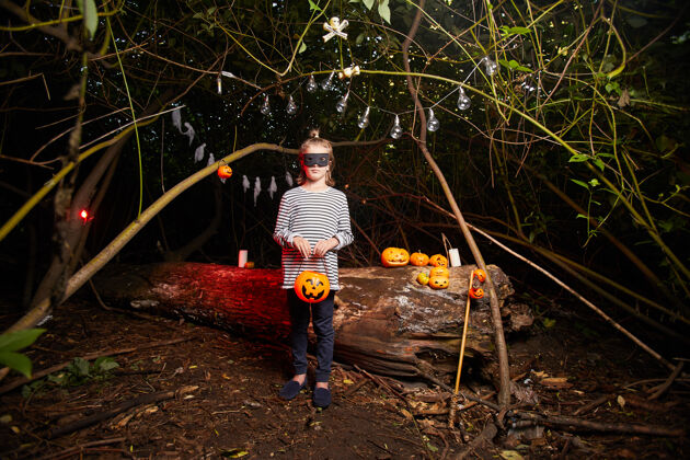 白天万圣节派对上穿着服装站在黑暗森林里的小女孩的画像叶子南瓜林地
