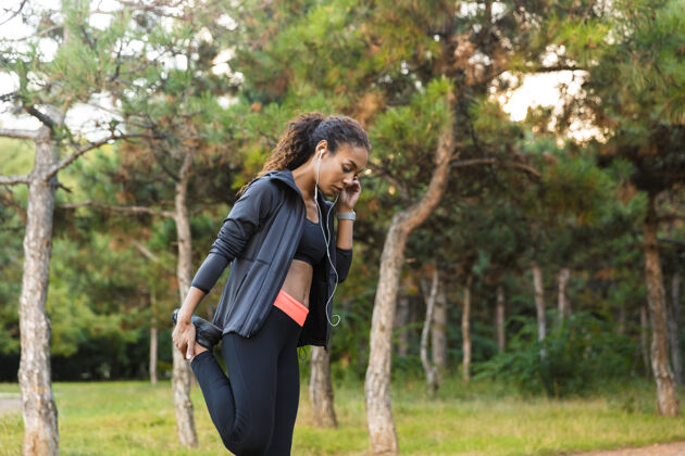精力充沛20多岁的美国黑人女性穿着黑色运动服 在绿色公园里伸展身体黑发耳机公园