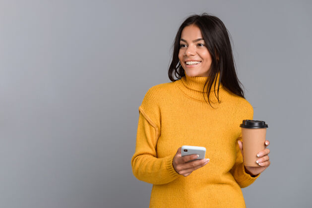 使用照片中 一位面带微笑的休闲年轻女子被隔离在灰色的墙上 手里拿着外卖咖啡 用着手机女孩温暖工作