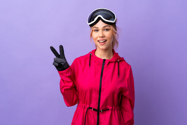 旅行滑雪女孩戴着滑雪板眼镜 脸上露出紫色的微笑和胜利的标志玻璃寒冷滑雪板