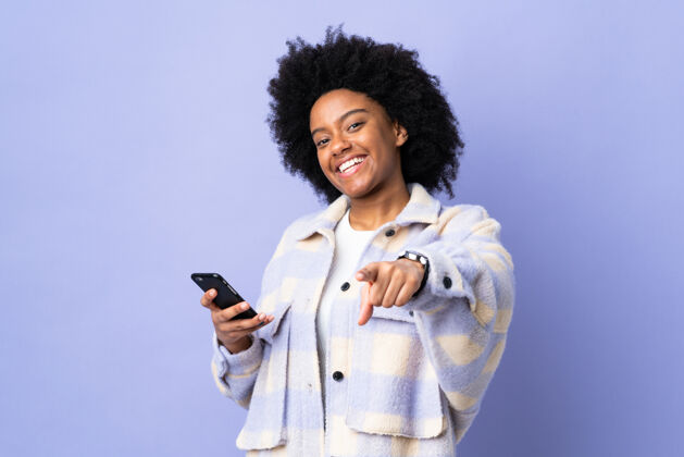 手机年轻的非洲裔美国妇女用手机孤立在紫色的背景点手指在你与一个自信的表情快乐颜色表情