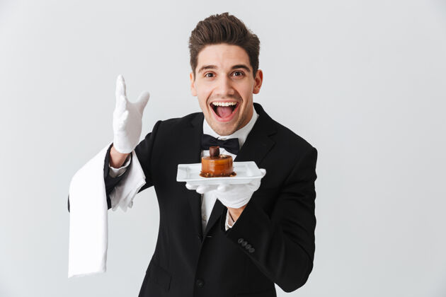 微笑穿着燕尾服的年轻帅哥 打着领结 拿着盘子 蛋糕隔着白墙工作人员单身食物