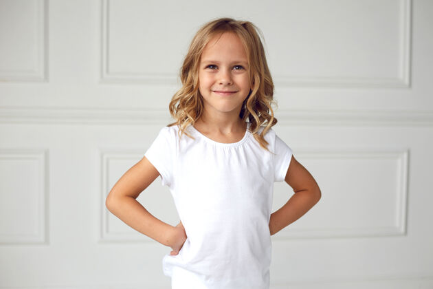 时尚正面肖像是一个小女孩微笑着看着镜头 穿着白色t恤 手放在腰上快乐可爱金发
