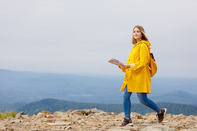 时髦穿着黄色雨衣的时髦女孩背着背包在山顶上看地图看绿色全景