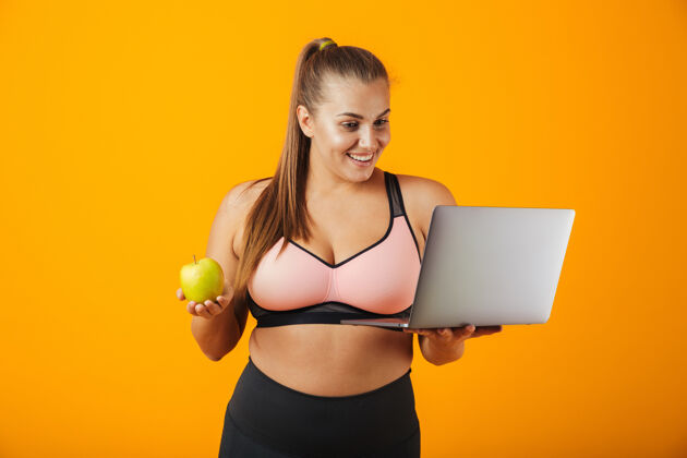 普拉提一个穿着运动服的快乐超重的年轻女子的肖像 孤立地站在黄色的墙上 用笔记本电脑 拿着绿色的苹果笔记本电脑通讯超重