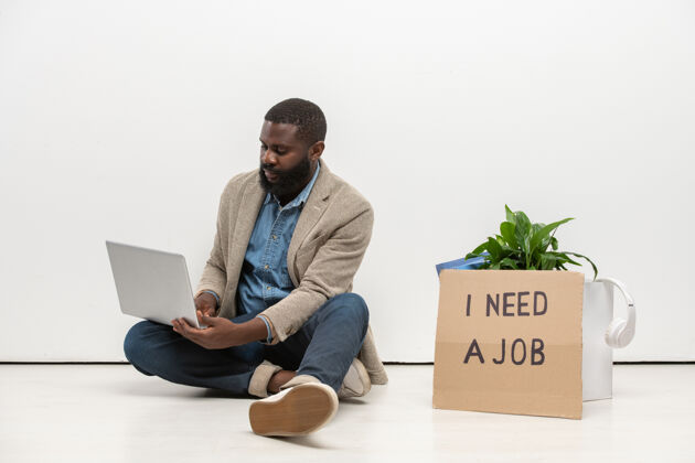 室内年轻的非洲失业者穿着休闲服坐在办公室墙边的地板上 用笔记本电脑在网上寻找新的空缺无线技术年轻人投票