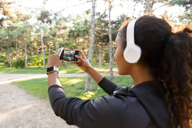 耳机一张20多岁的非洲裔美国妇女的肖像 她穿着黑色运动服 戴着耳机 在绿色公园散步时用手机自拍身体非洲户外