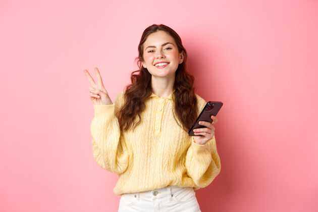 成人科技和网上购物快乐的年轻女子微笑着高兴 手持智能手机 显示v型标志 看着相机 站在粉红色的墙上年轻在线微笑