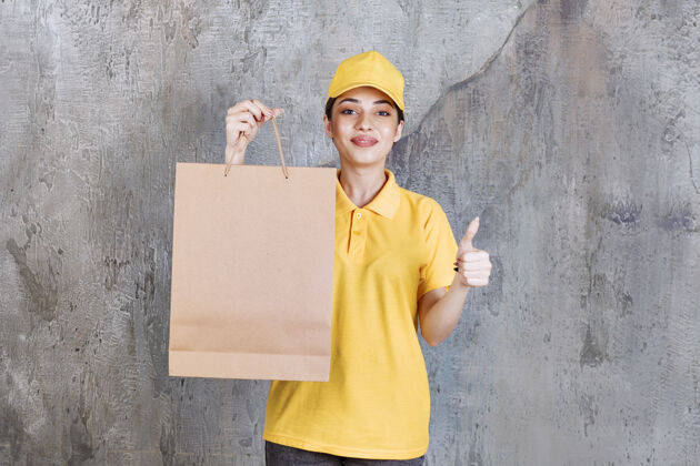 年轻人身着黄色制服的女服务人员手持购物袋 并显示积极的手势交易员工送货
