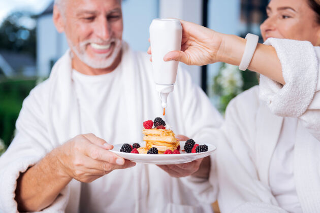 水果蜂蜜煎饼美丽的妻子戴着白色智能手表在手上使用蜂蜜煎饼 而与丈夫吃早餐咖啡放松果汁